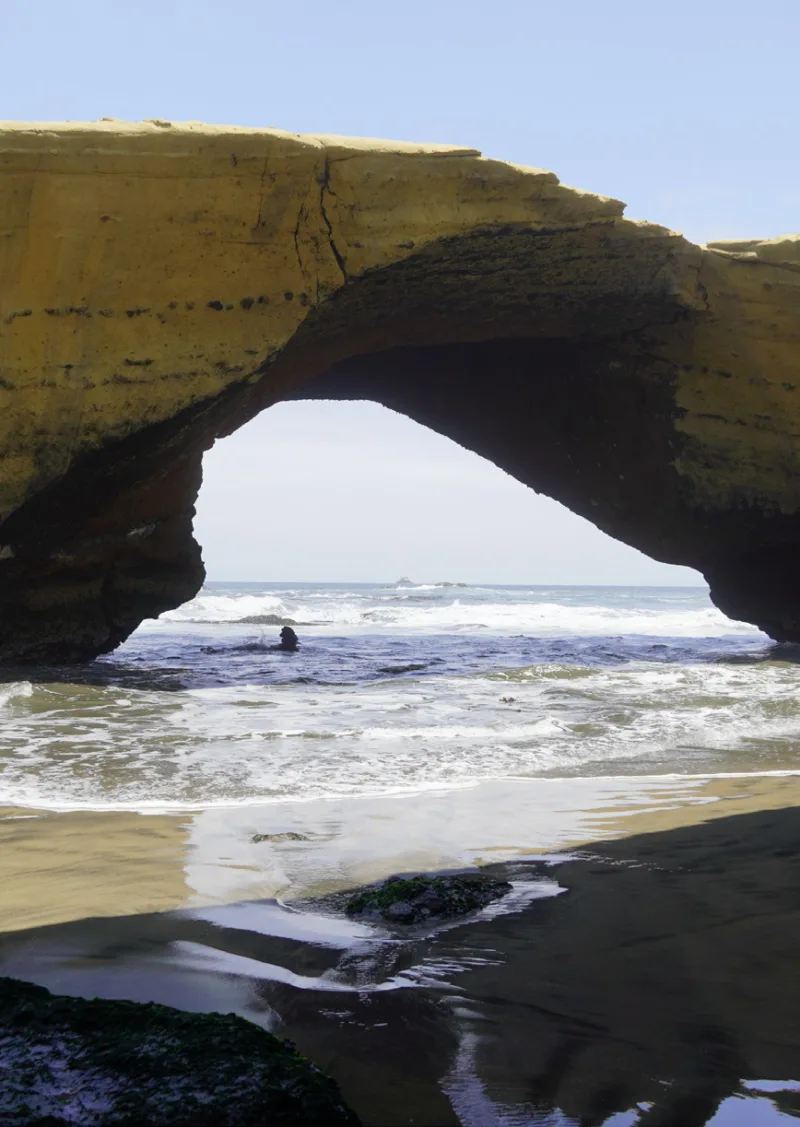 Gran arco natural de roca junto al mar, con olas y orilla de arena.