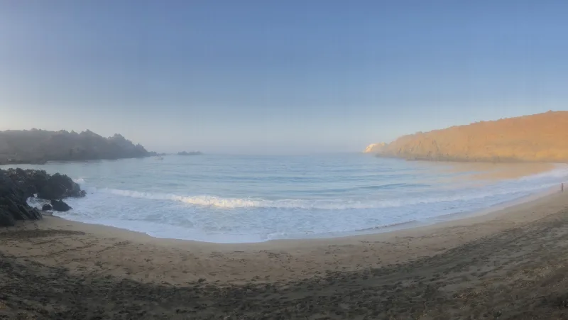 Ensenada de arena rodeada de promontorios rocosos y mar al fondo.