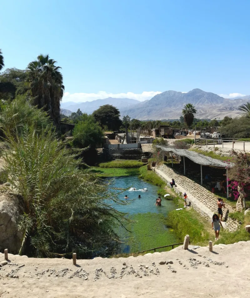 Poza de agua de tono verdoso con personas, vegetación y construcciones alrededor, y cerros al fondo.