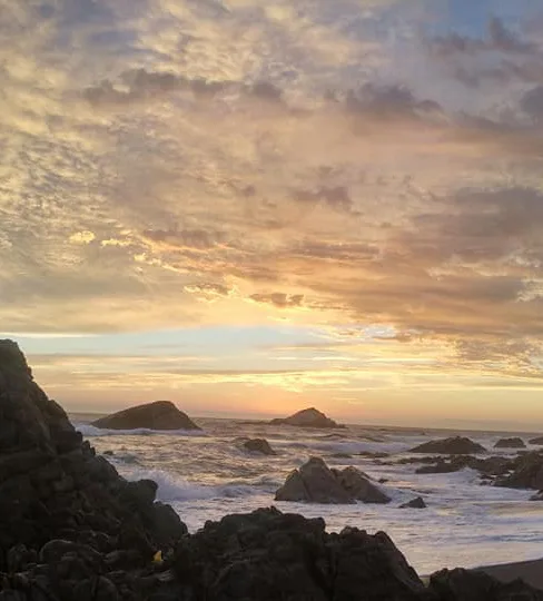 Costa de rocas oscuras, espuma de las olas y un cielo de tonos cálidos al atardecer.