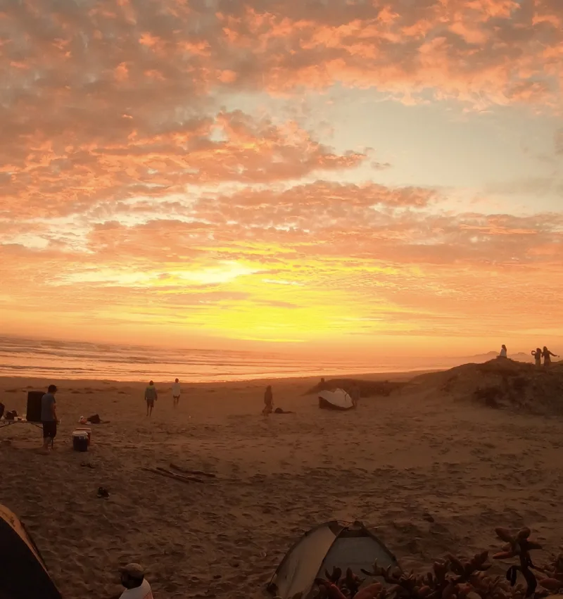 Atardecer de cielo anaranjado sobre una playa con personas y tiendas en la arena.